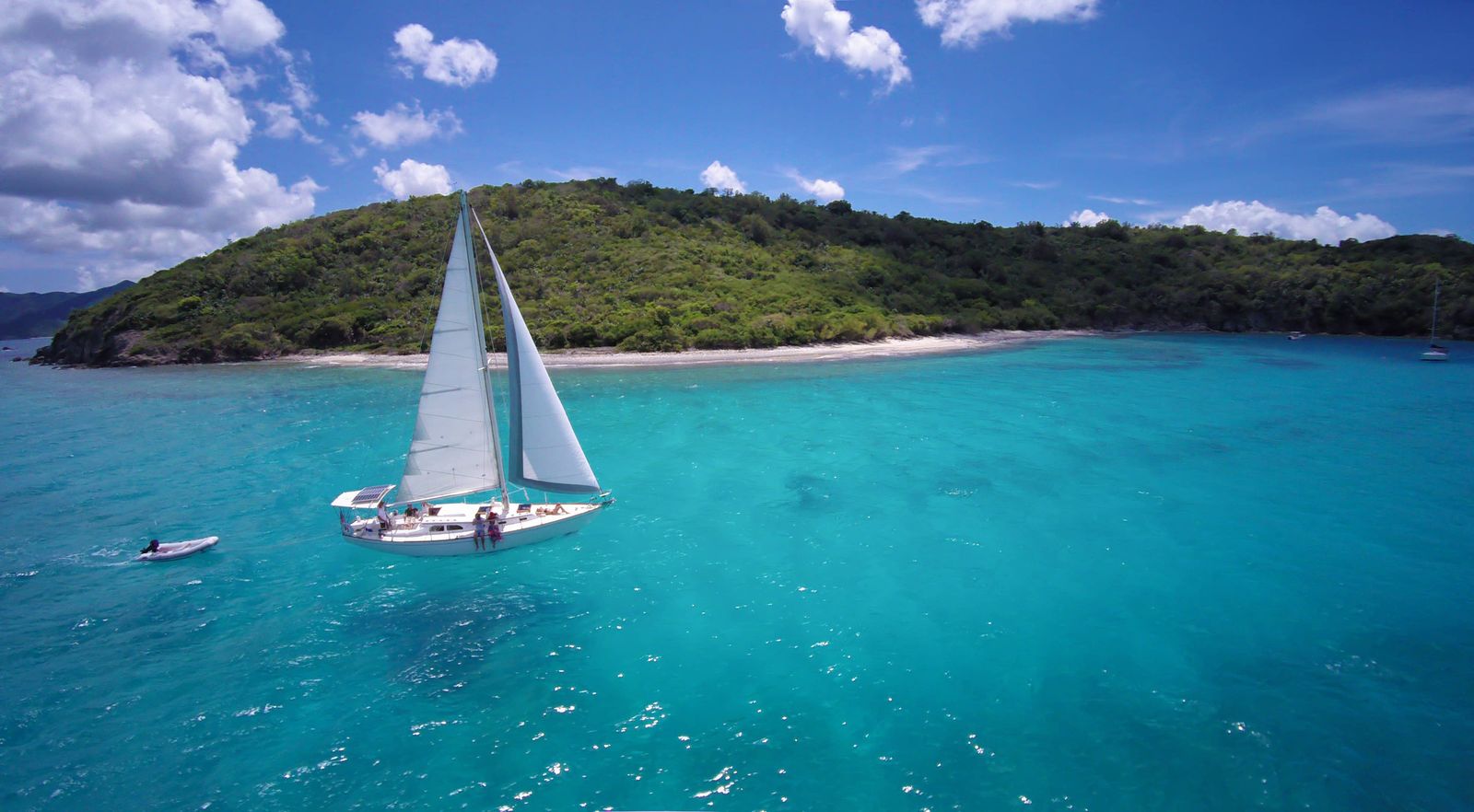 Sailing yacht under sail with its crew off a green island, tender in tow