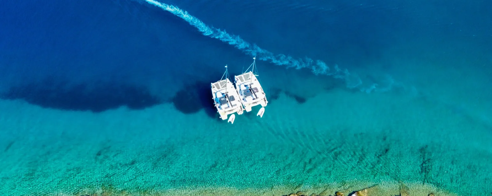 Two charter catamarans anchored off a rocky shore in clear turquoise water