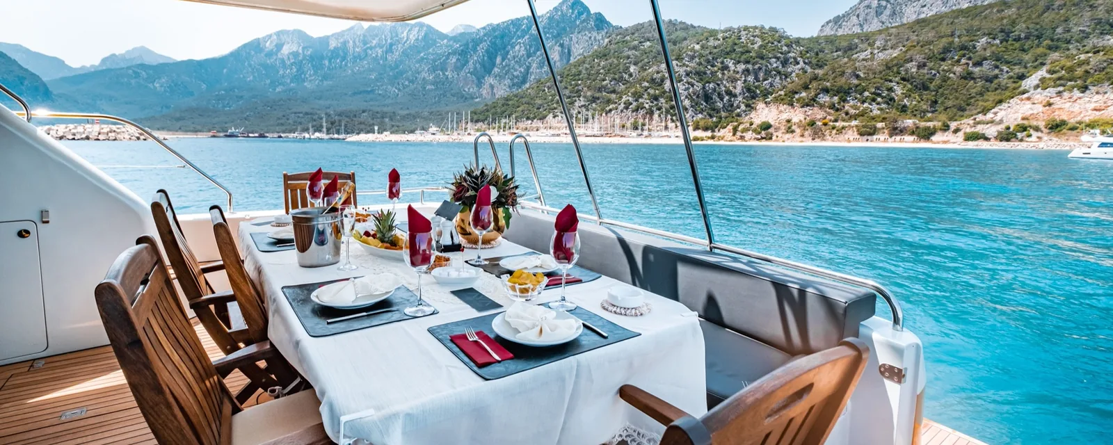 Table set for lunch on the aft deck of a crewed motor yacht at anchor, with mountains along the coast