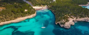 Aerial view of a sailing yacht anchored in a turquoise cala lined with pine forest and limestone cliffs