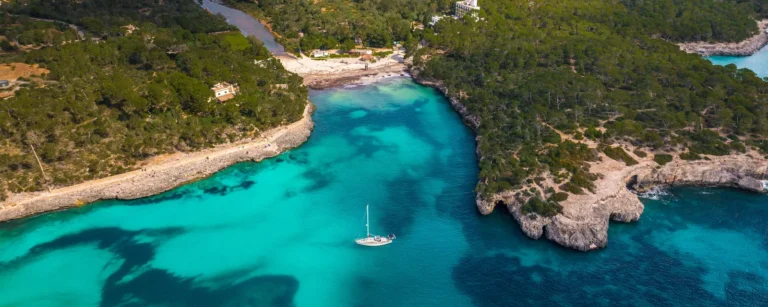 Aerial view of a sailing yacht anchored in a turquoise cala lined with pine forest and limestone cliffs