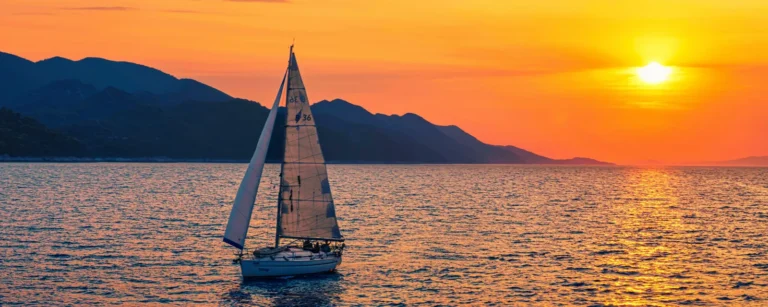 A sailing yacht under sail at sunset along a mountainous coast