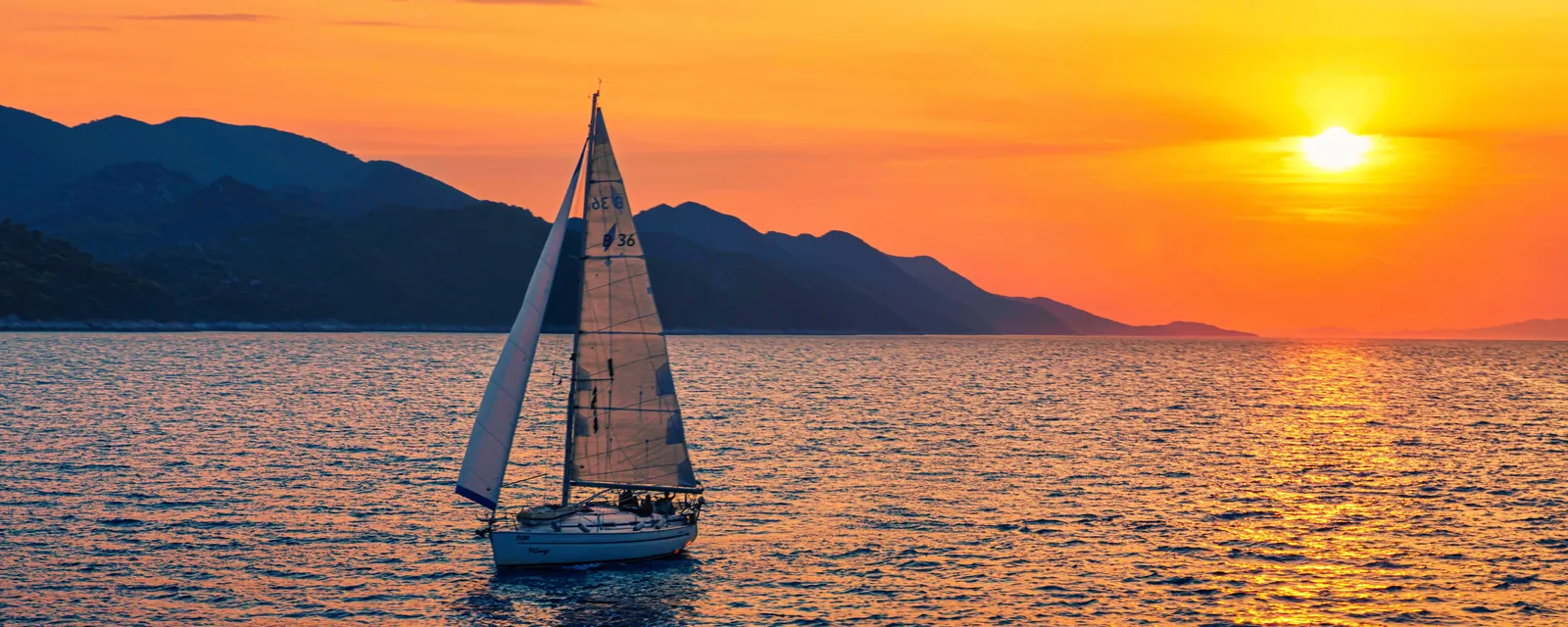 A sailing yacht under sail at sunset along a mountainous coast