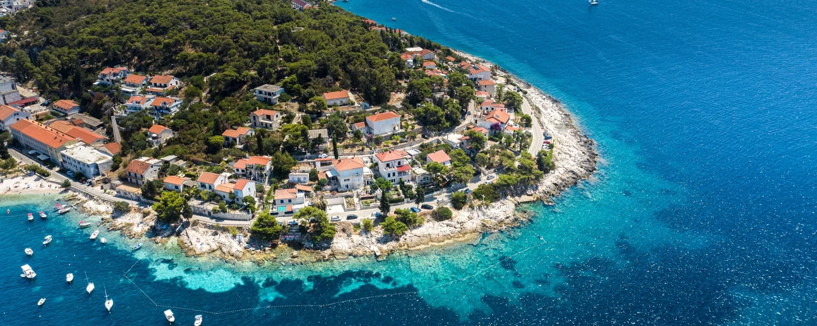 Aerial view of an island village on a wooded headland, with boats anchored in clear turquoise water