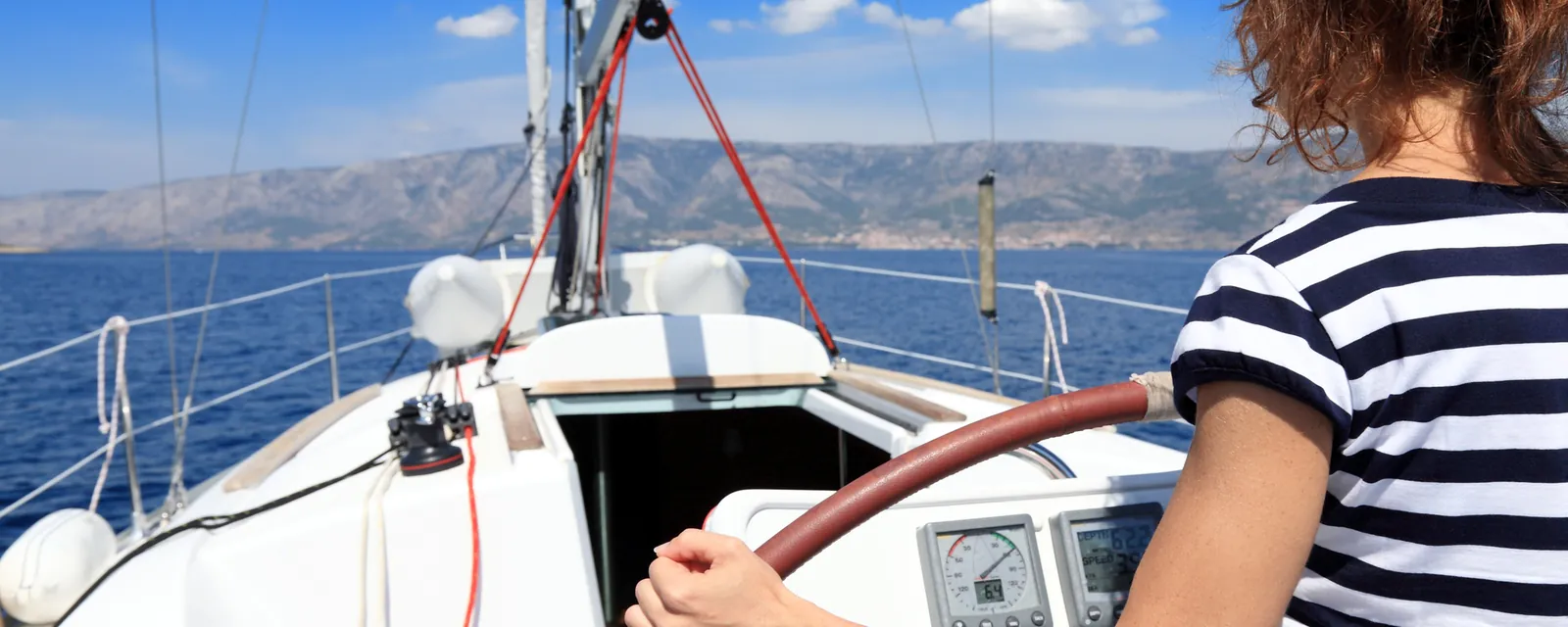 A woman at the helm of a sailing yacht with an island on the horizon