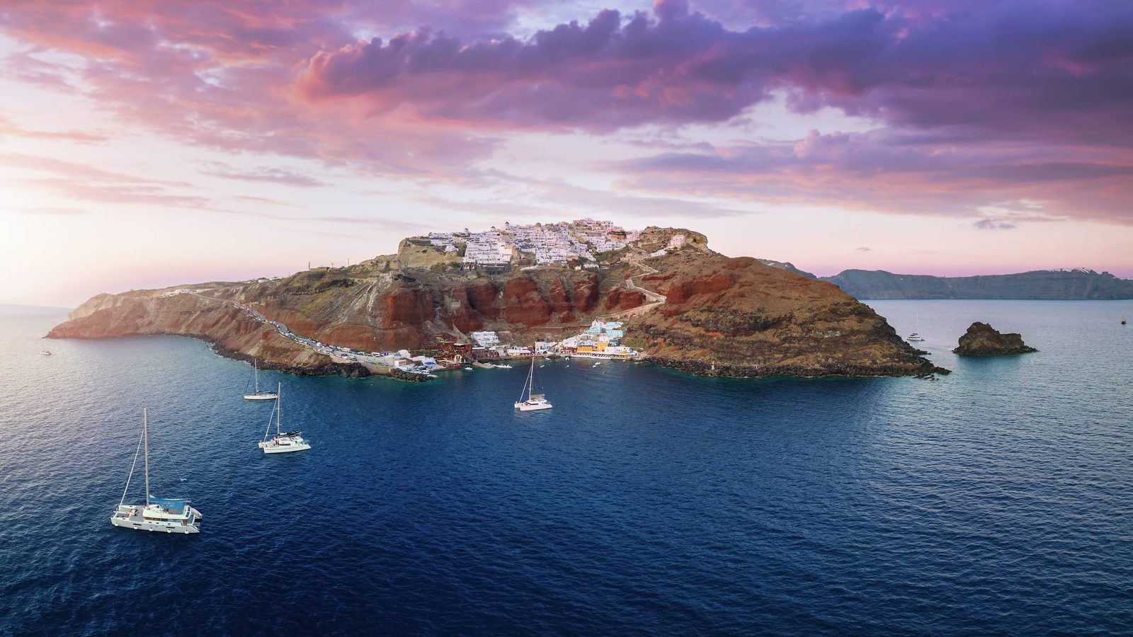 Sailing yachts and catamarans anchored below Oia and Ammoudi Bay, Santorini, at sunset